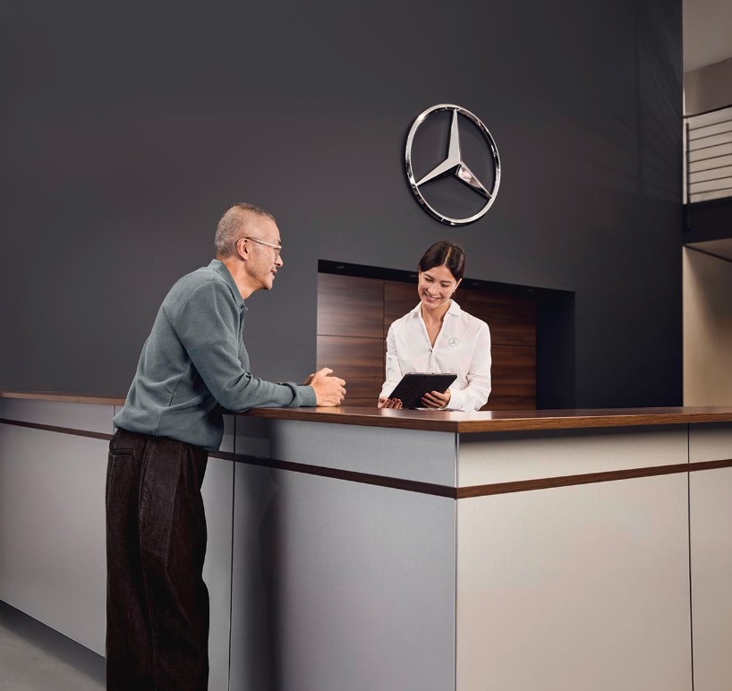 Beratung zu Mercedes-Benz Originalteilen Empfangsbereich der Serviceabteilung eines Mercedes-Benz-Händlers, wo ein Kunde mit einem Serviceberater am Empfang die Einzelheiten der Fahrzeugwartung bespricht.