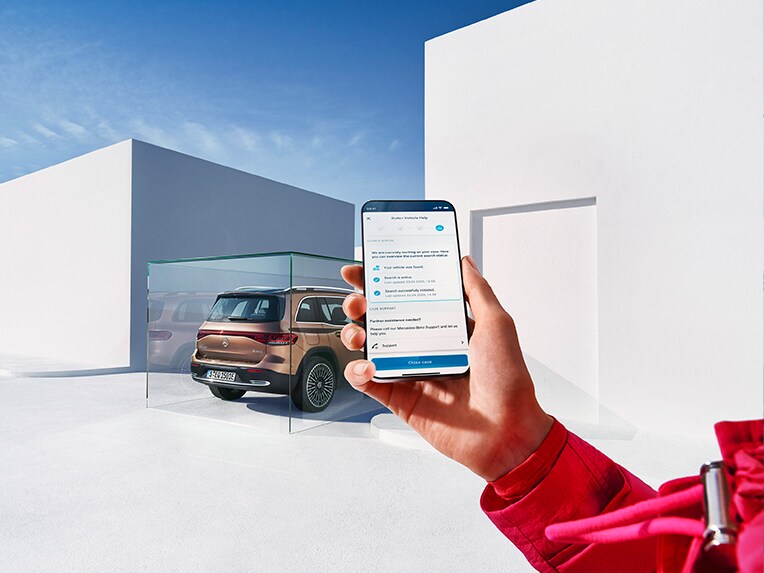 Eine Person hält ein Mobiltelefon und nutzt die Mercedes-Benz App. Im Hintergrund parkt ein EQB.