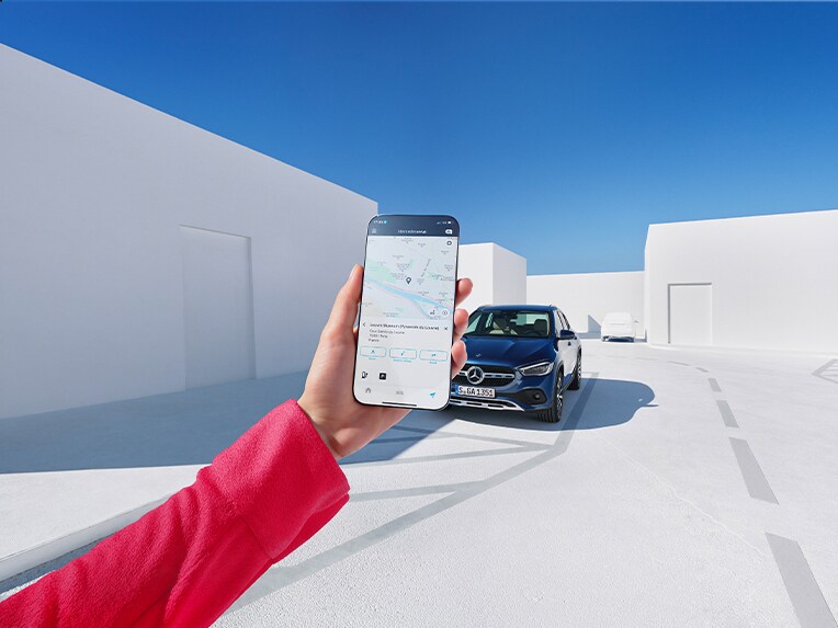 Eine Frau informiert sich per Mercedes me App auf ihrem Smartphone und den Digitalen Extras zum Thema Navigation, wie das Wetter am Ankunftsort wird.