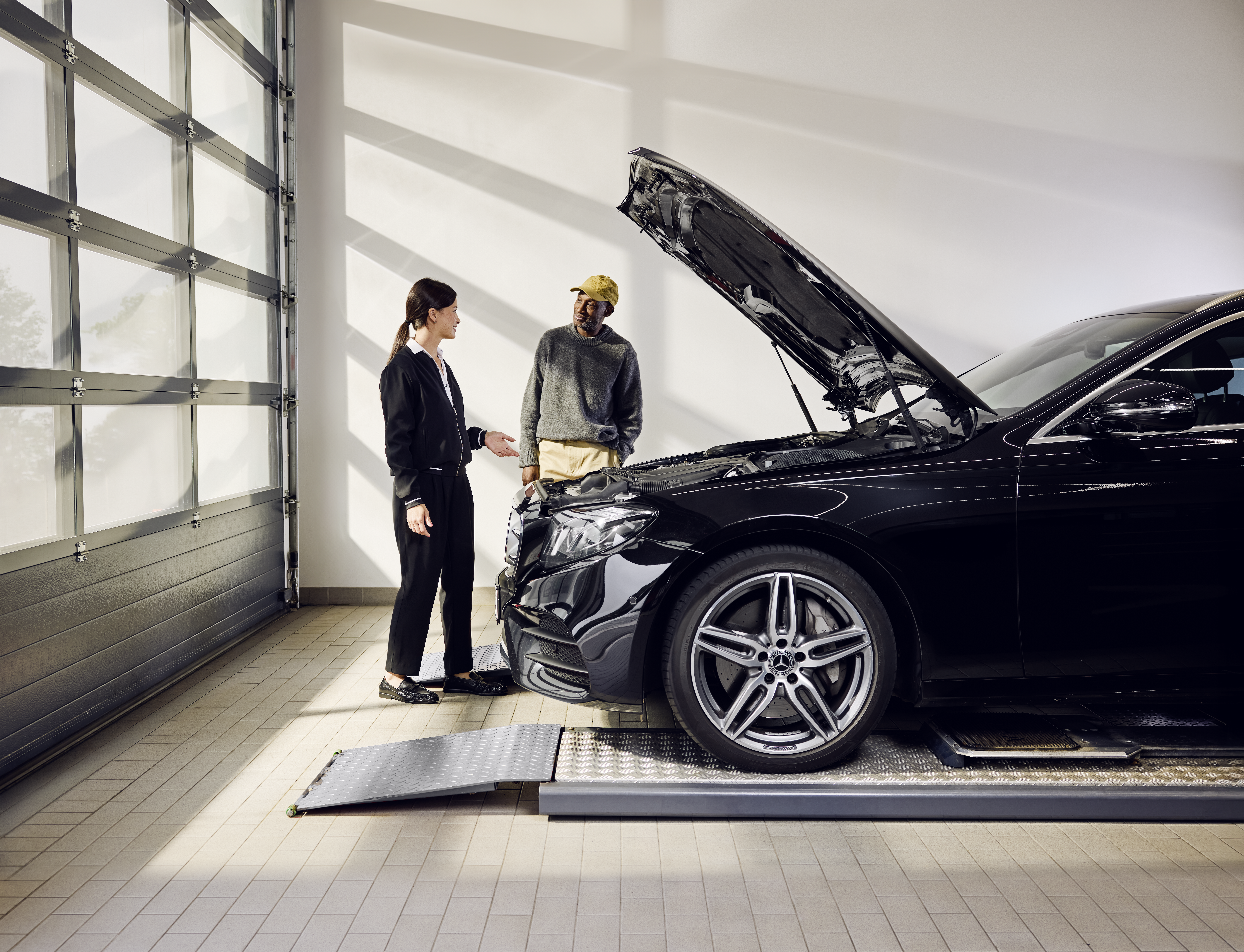 Austausch mit Originalteilen von Mercedes-Benz Ein Mercedes-Benz-Fahrzeug, das während eines fachgerechten Austauschs von Originalteilen in einer Werkstatt auf einer Servicerampe steht und dessen Motorhaube geöffnet ist.