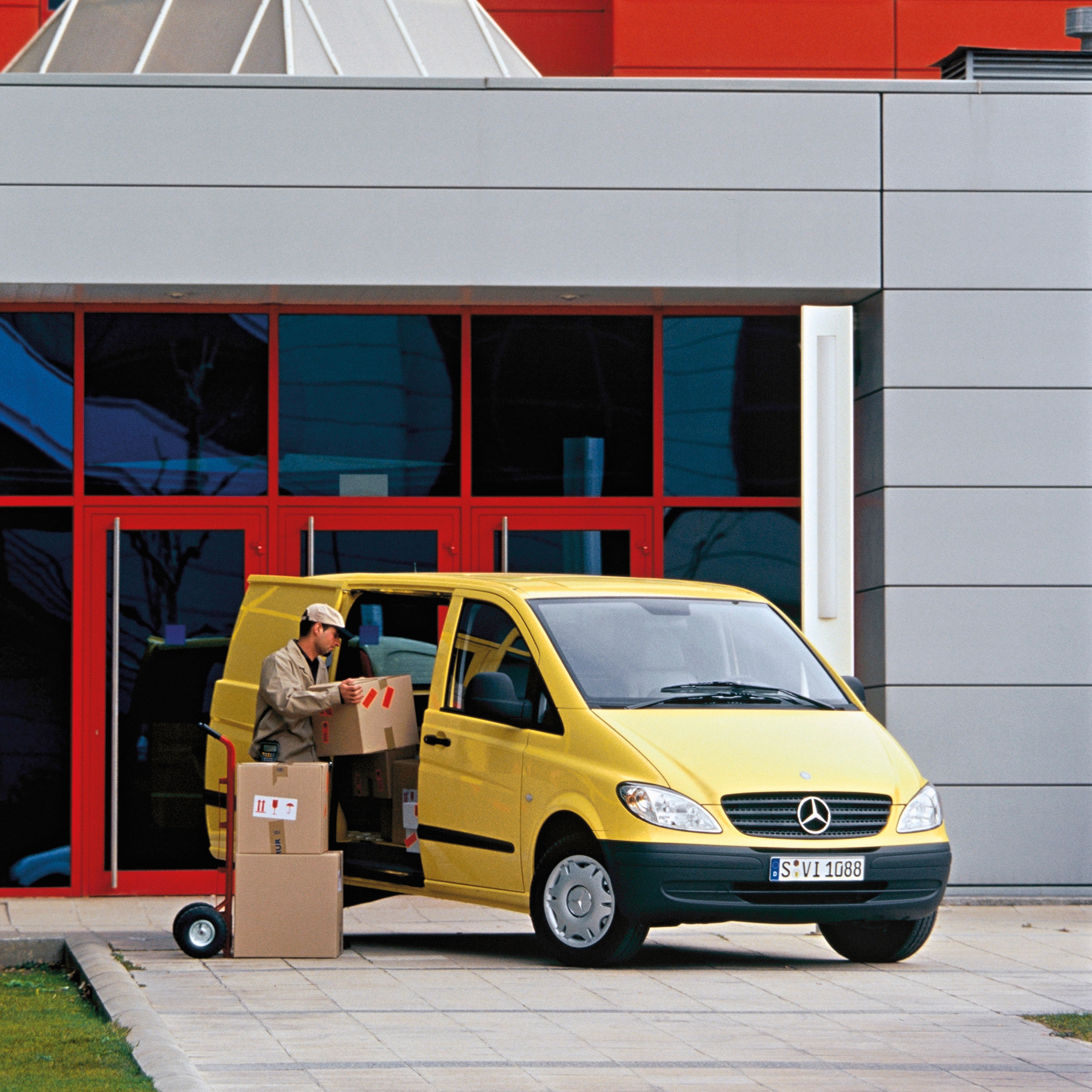 Die zweite Generation des Mercedes-Benz Vito | Geschichte der Nutzfahrzeuge | Mercedes-Benz Ein gelber Mercedes-Benz Vito, der vor dem Gebäude geparkt ist, während ein Arbeiter Pakete auslädt
