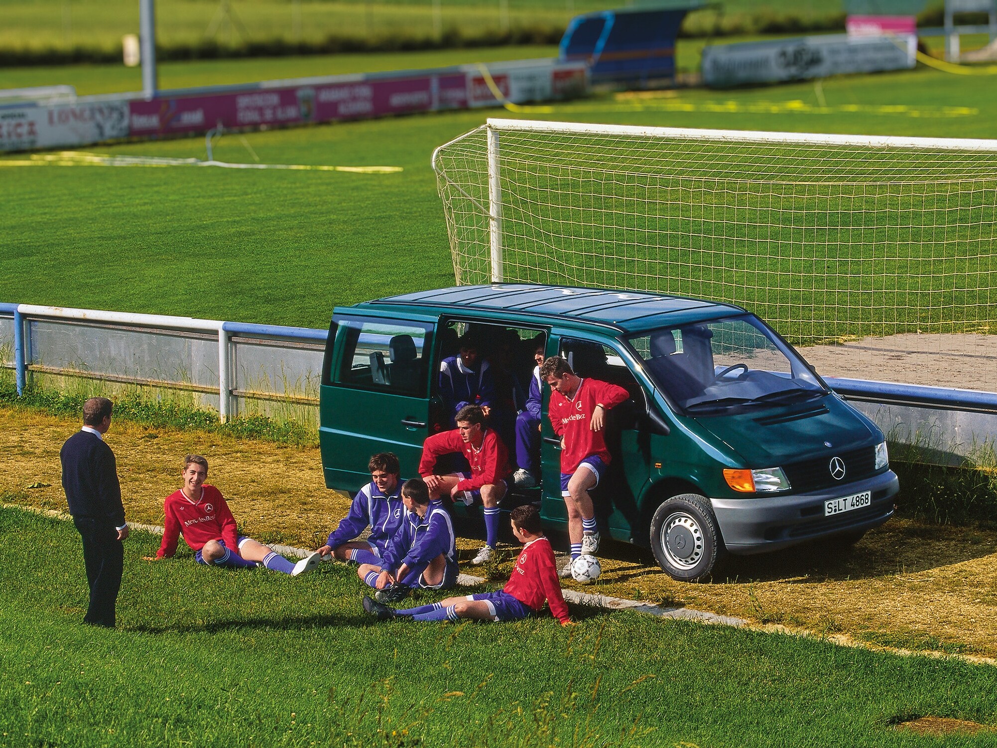 Ein Vito der ersten Generation, der neben einem Fußballplatz geparkt ist, mit Spielern der Mannschaft in der Nähe