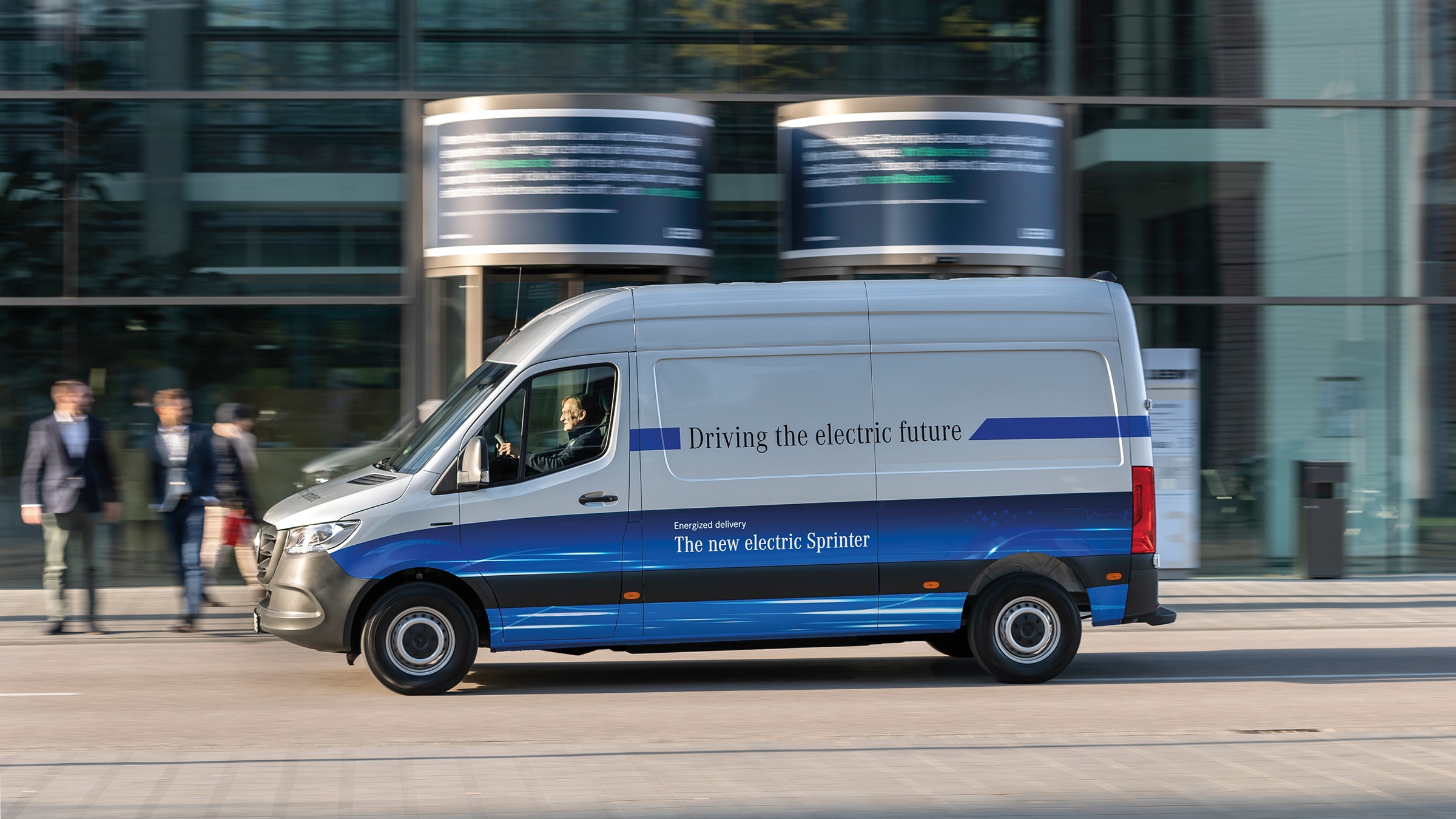 Der eSprinter Kastenwagen der zweiten Generation | Geschichte der Nutzfahrzeuge | Mercedes-Benz Ein Mercedes-Benz eSprinter Kastenwagen fährt durch eine moderne Stadtstraße