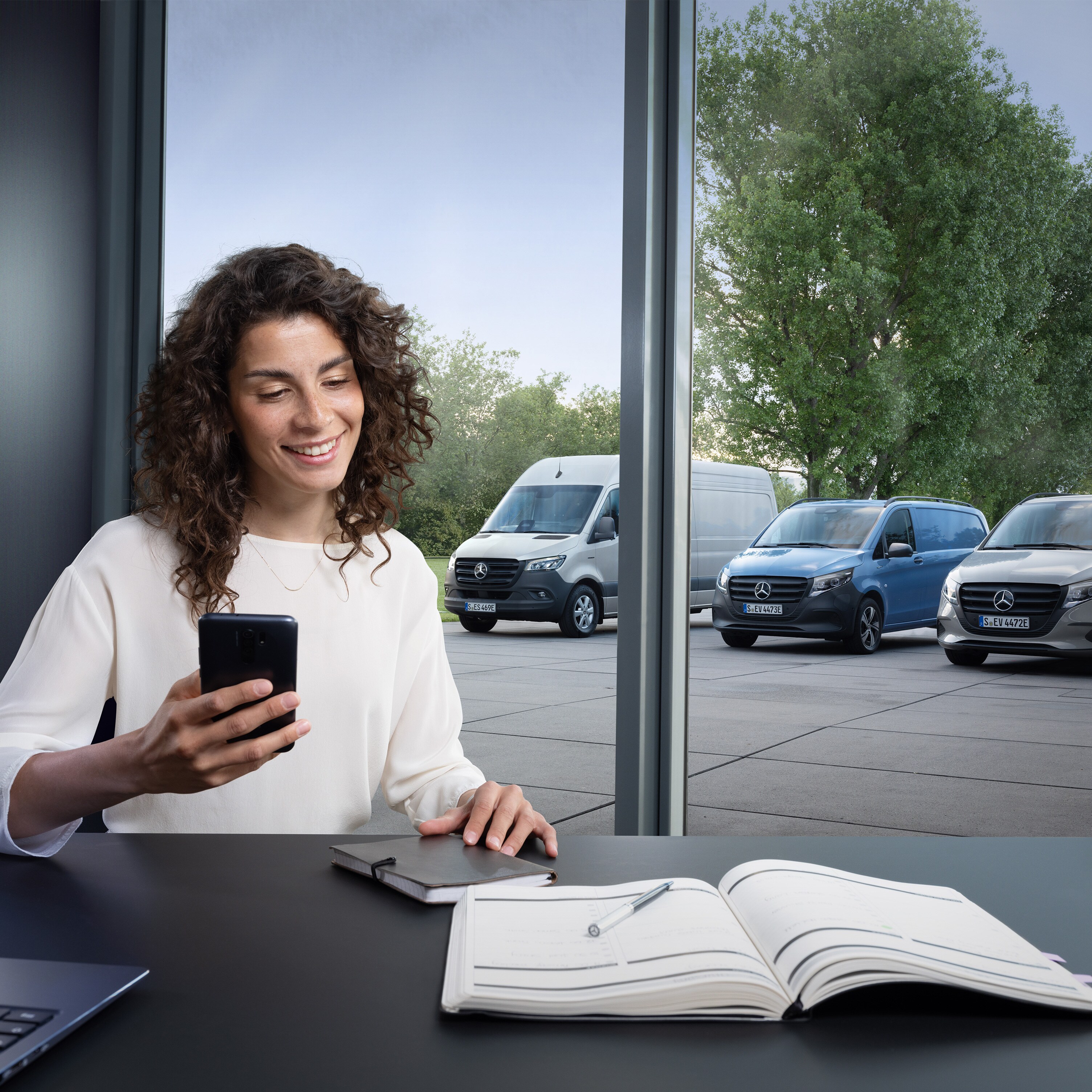 Eine Frau plant auf ihrem Smartphone vor einer Reihe von Mercedes-Benz Transportern im Hintergrund.