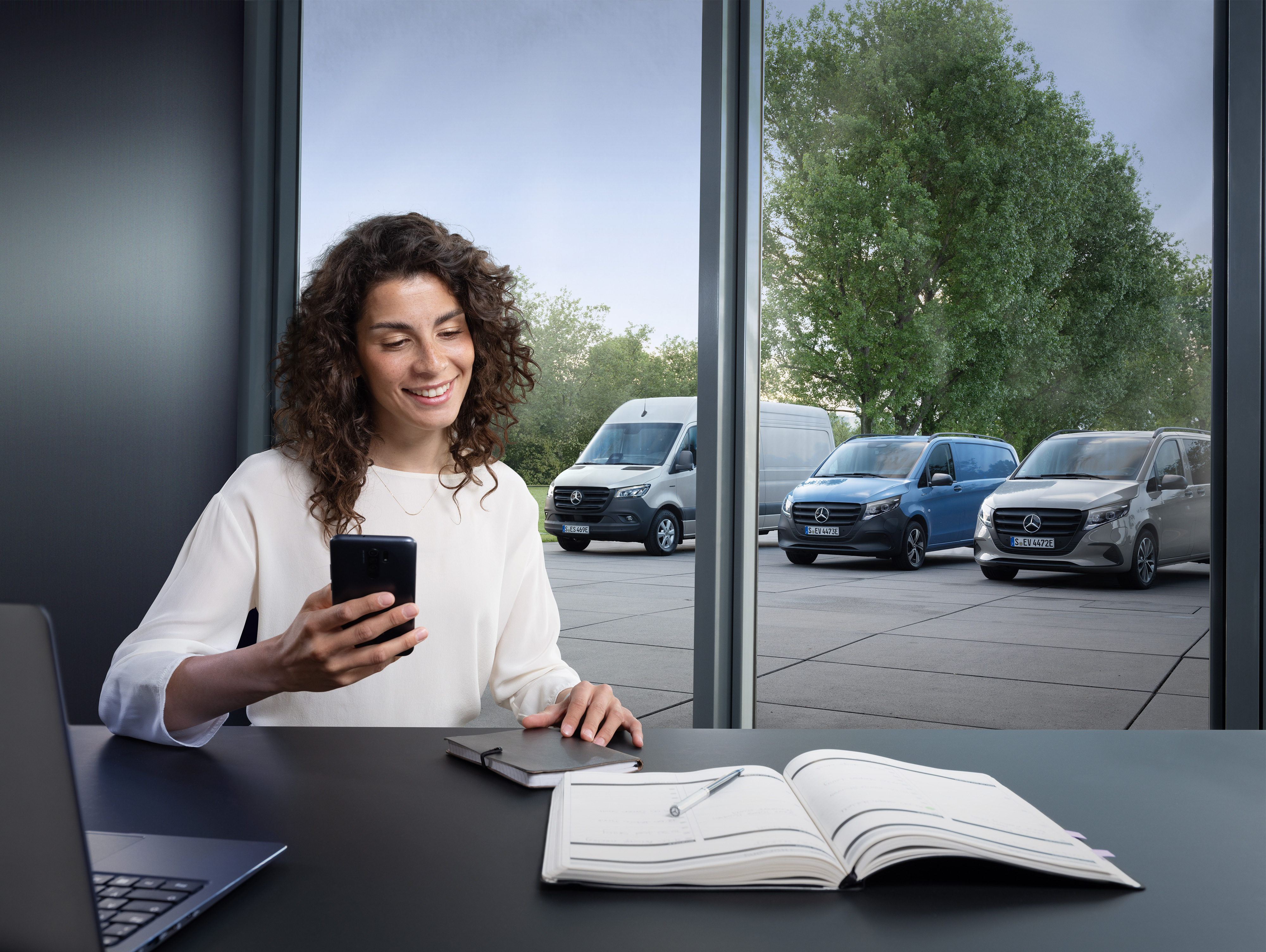 Mercedes-Benz | Van ServiceCare | Terminvereinbarung Eine Frau plant auf ihrem Smartphone vor einer Reihe von Mercedes-Benz Transportern im Hintergrund.